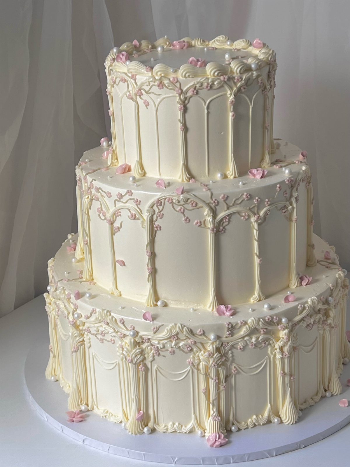 Three-tiered cream wedding cake with architectural frosting, pink flowers, and pearl accents.