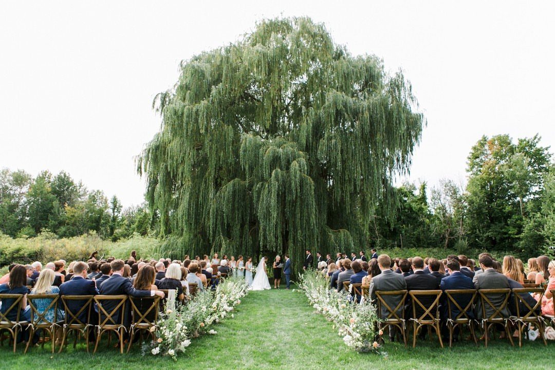 Fantasy Wedding Venue Ideas: 11 Storybook Settings 9 Wedding ceremony under a large weeping willow tree with guests in wooden chairs.