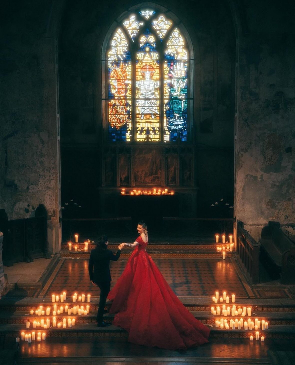 8 Gothic Romance Wedding Ideas for a Dark, Moody Day 1 Couple in red gown and black suit standing in a gothic cathedral with candles.