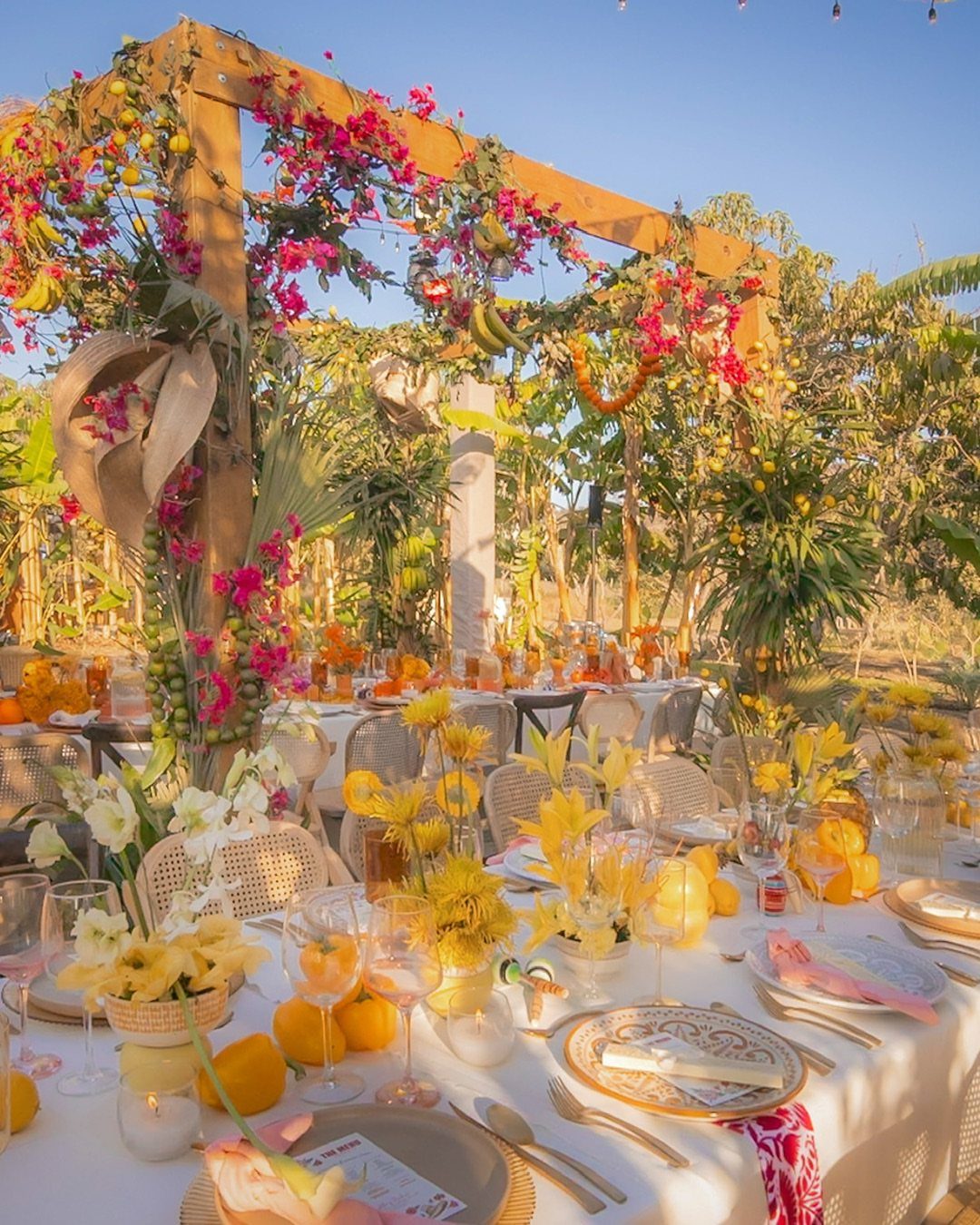 Wooden wedding arbor with pink flowers and hanging bananas over a sunny table.