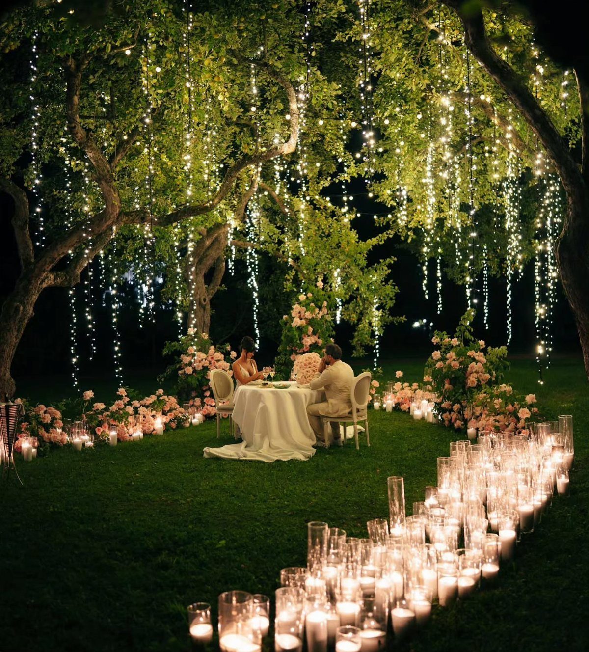 Romantic outdoor dinner under a tree with fairy lights and candlelit pathways.