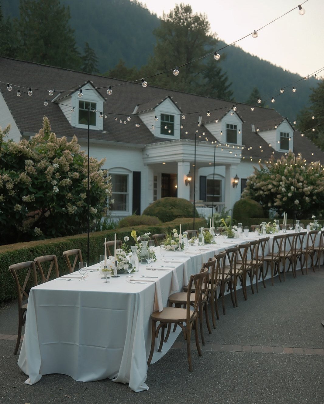 Long white wedding table with wooden chairs outside a white historic estate house.