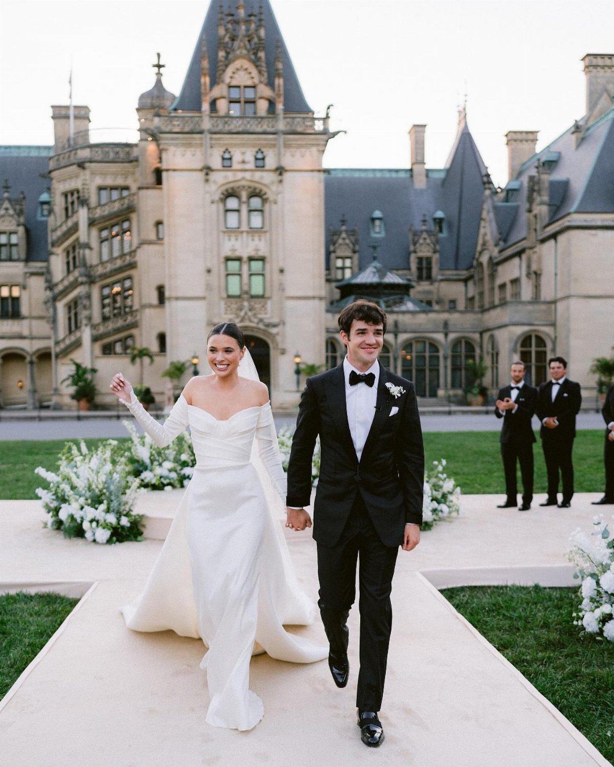 Newlywed couple walking before a grand historic stone mansion with ornate architecture and gardens.