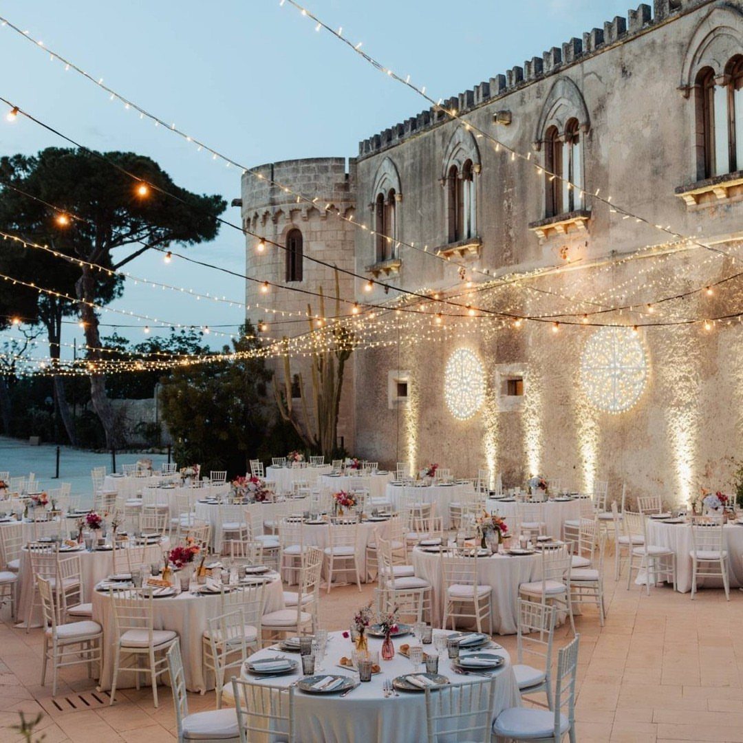 Outdoor wedding reception with white tables and fairy lights beside a historic stone building.