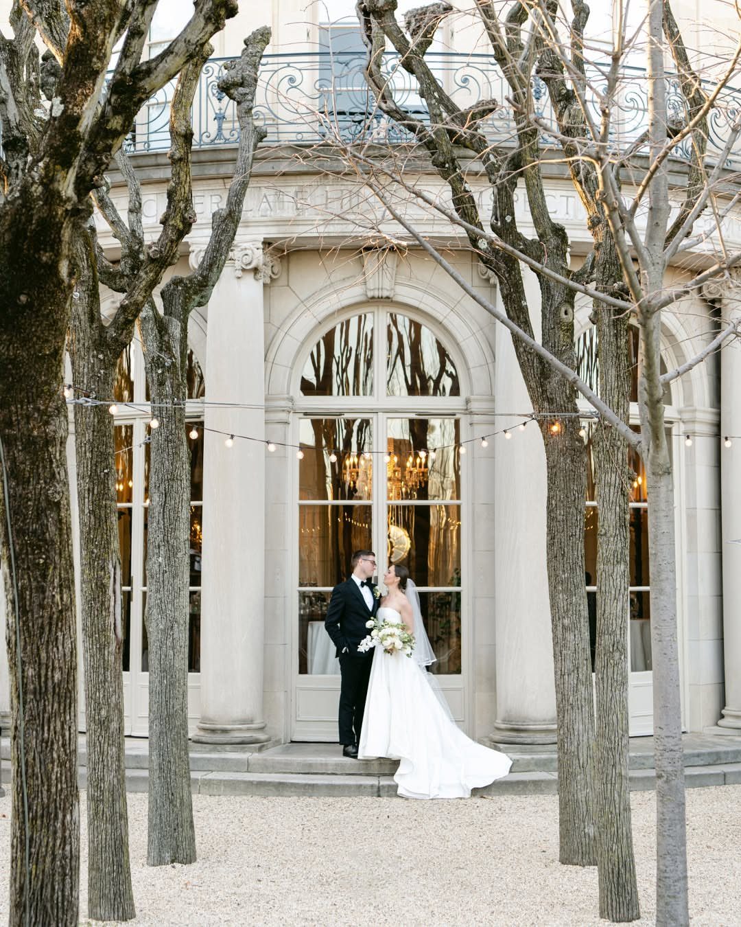 Bride and groom posing before a grand building with arched windows and bare trees.