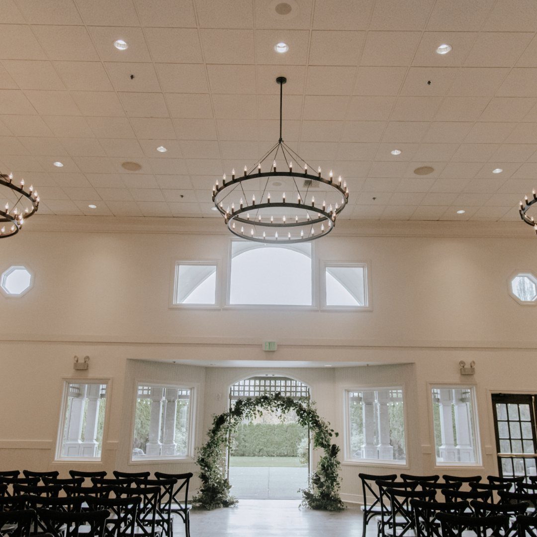 Indoor wedding ceremony space with black chairs, a floral arch, and large circular chandeliers.