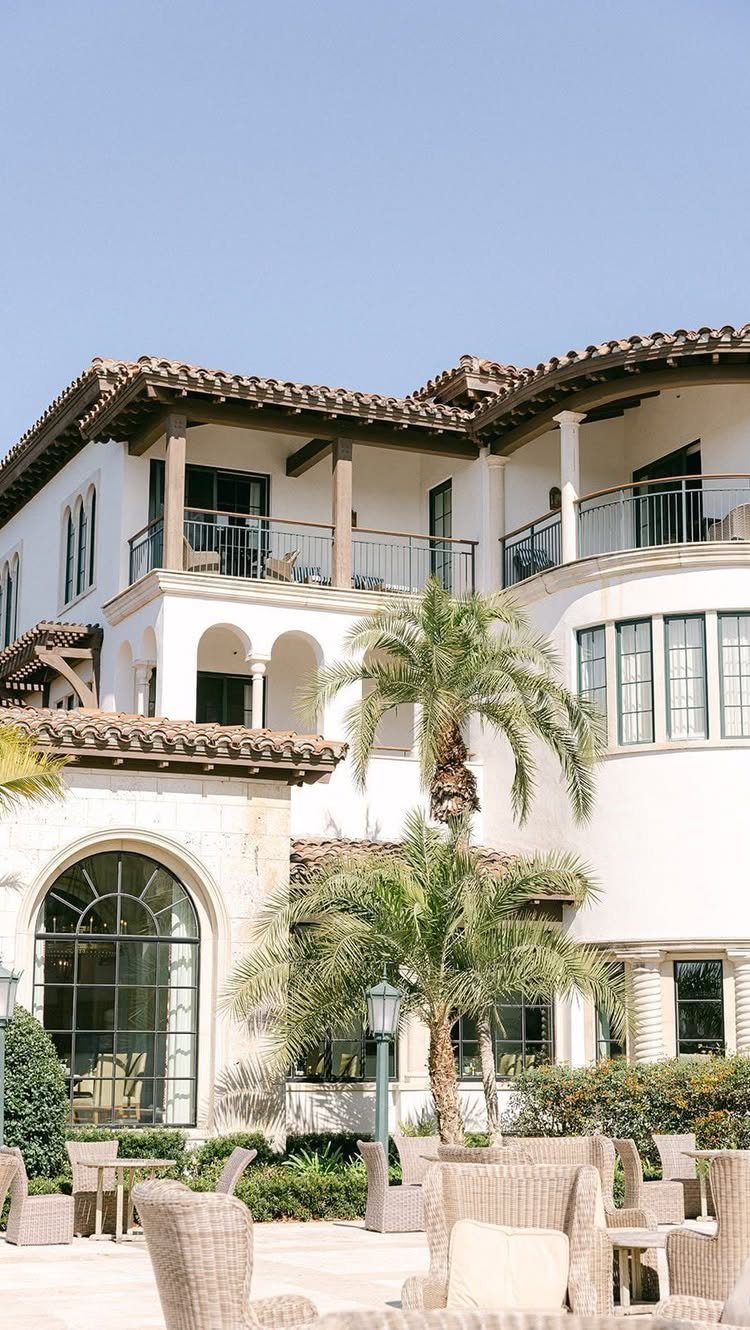 White Mediterranean villa with terracotta roof, palm trees, and wicker patio furniture outdoors.