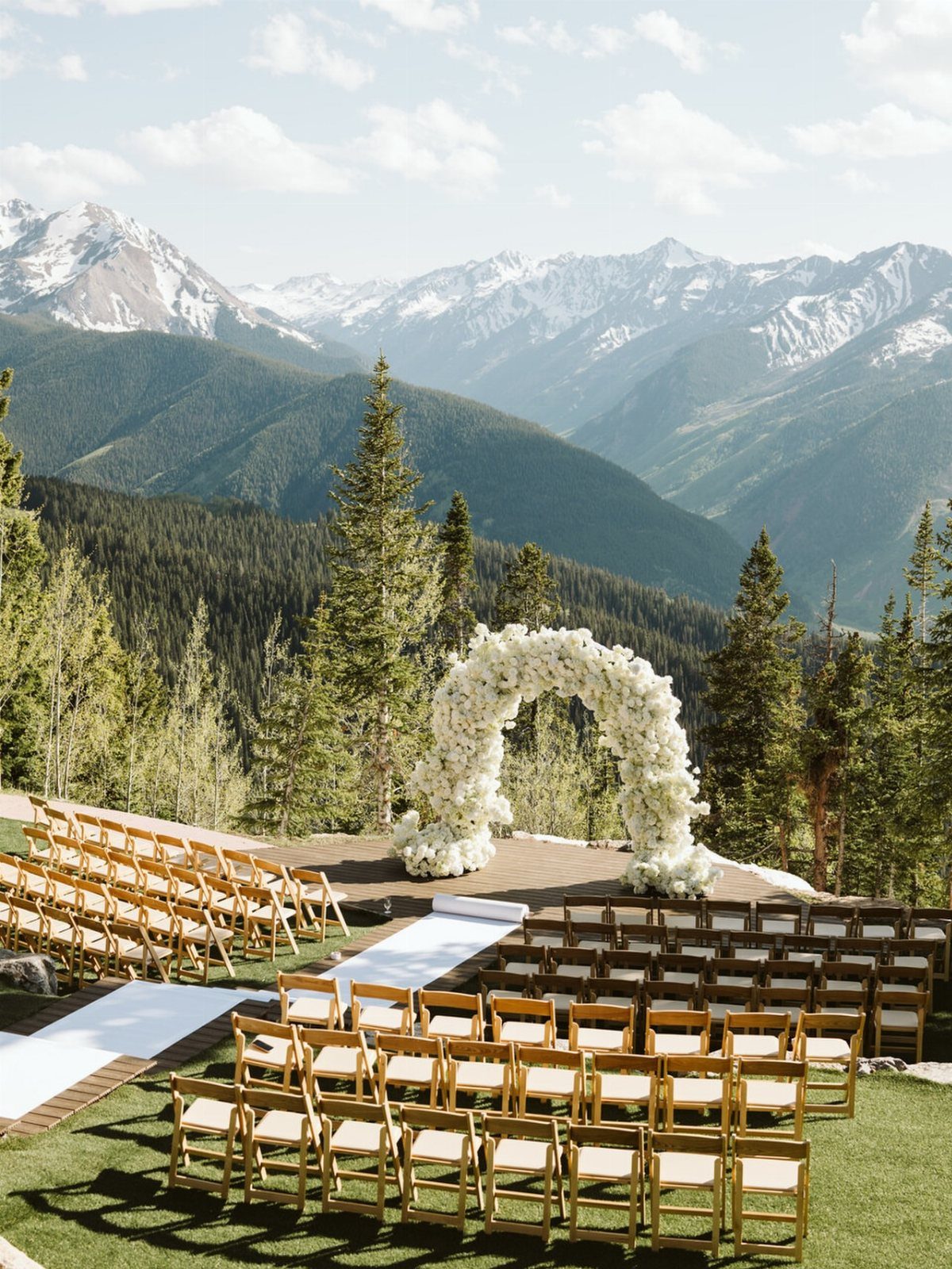 Outdoor wedding ceremony with a white floral arch overlooking snow-capped mountain peaks.