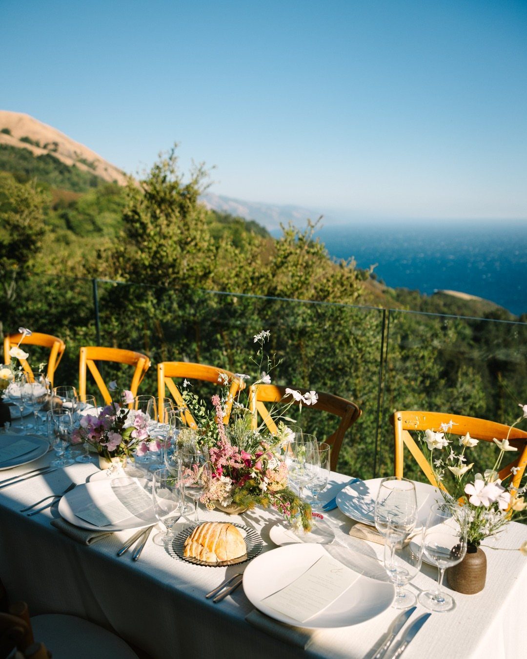 Dining table with white linens and yellow chairs overlooking a coastal hillside and ocean.