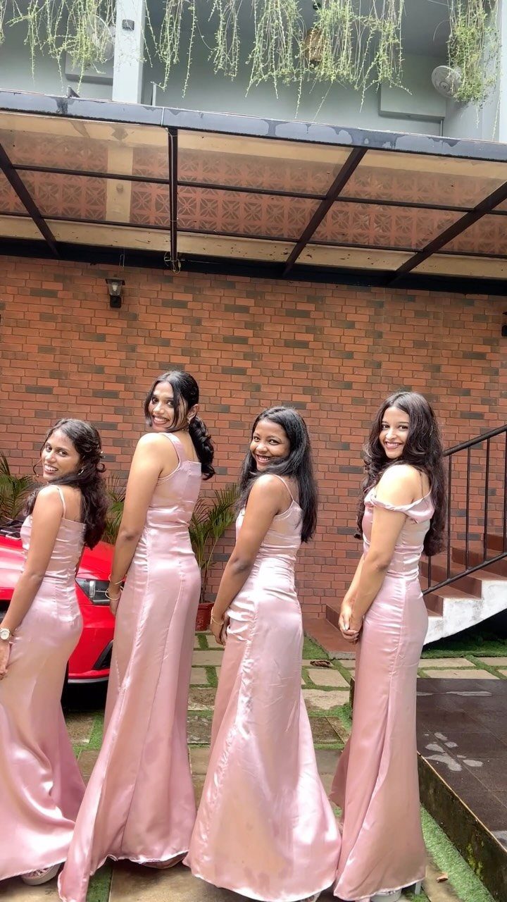 Four women posing in shimmering light pink satin bridesmaid dresses against a brick wall.