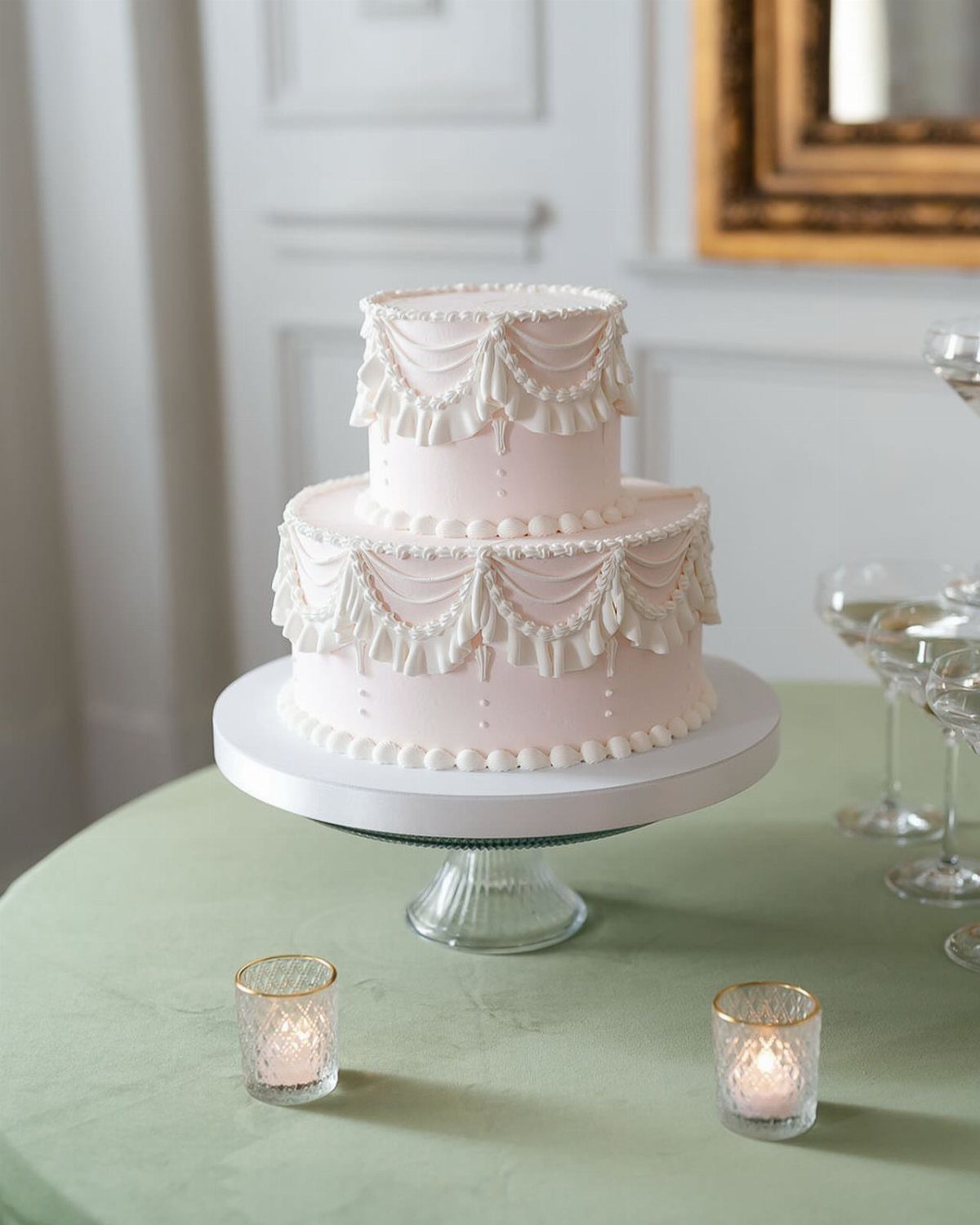 Two-tier light pink wedding cake with white frosting details on a sage green table.