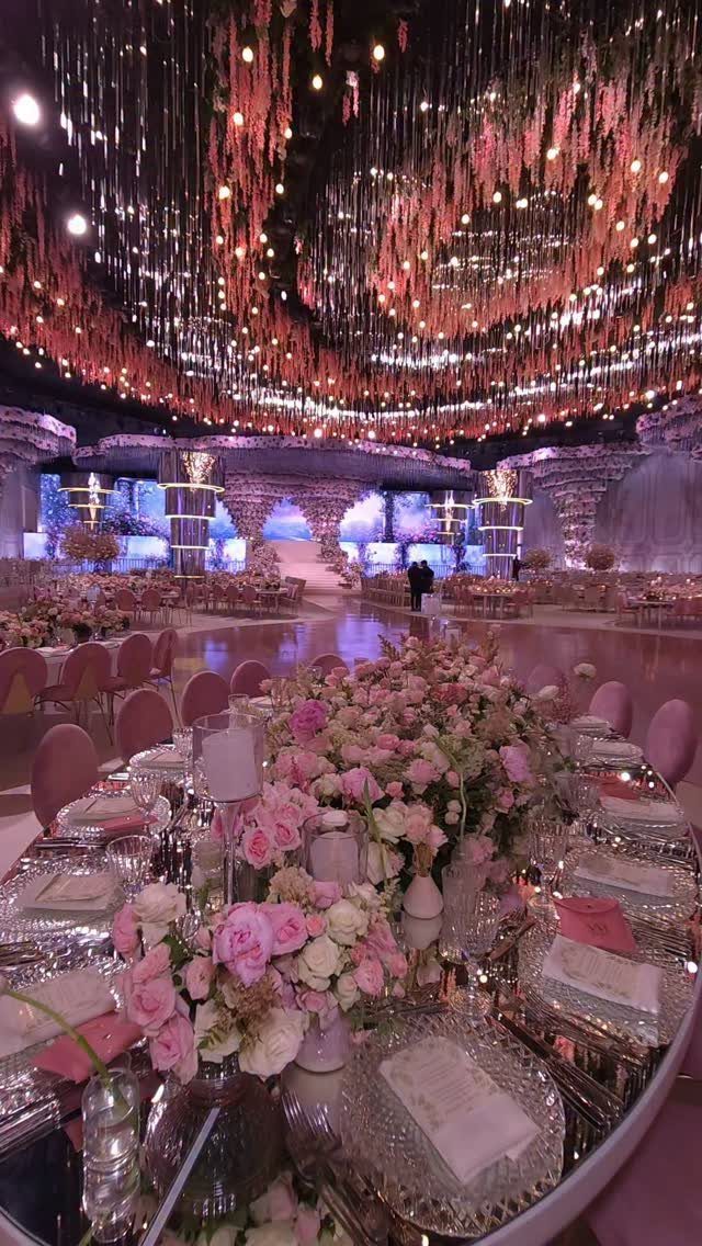 Lavish wedding reception hall with light pink floral centerpieces and hanging ceiling lights.