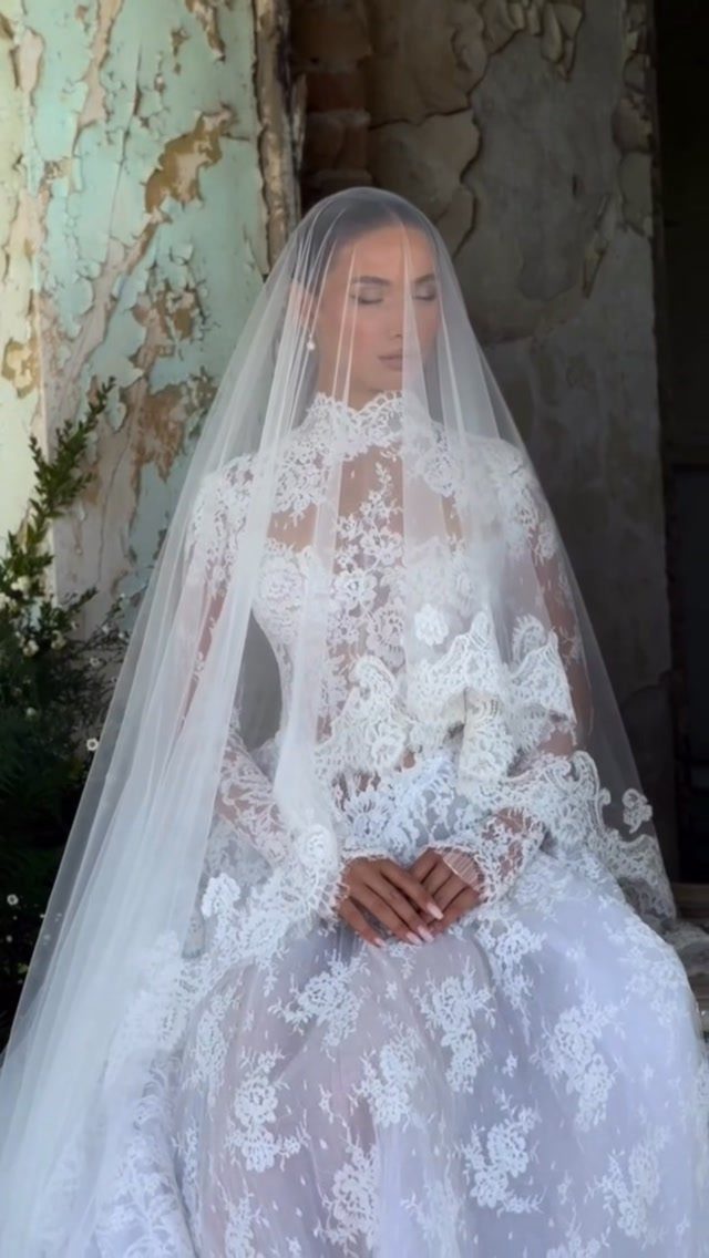 Woman in a long white lace wedding dress and sheer veil posing by a wall.