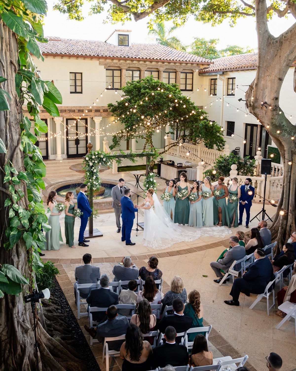 Wedding ceremony in a Mediterranean courtyard with floral arch, banyan trees, and guests.