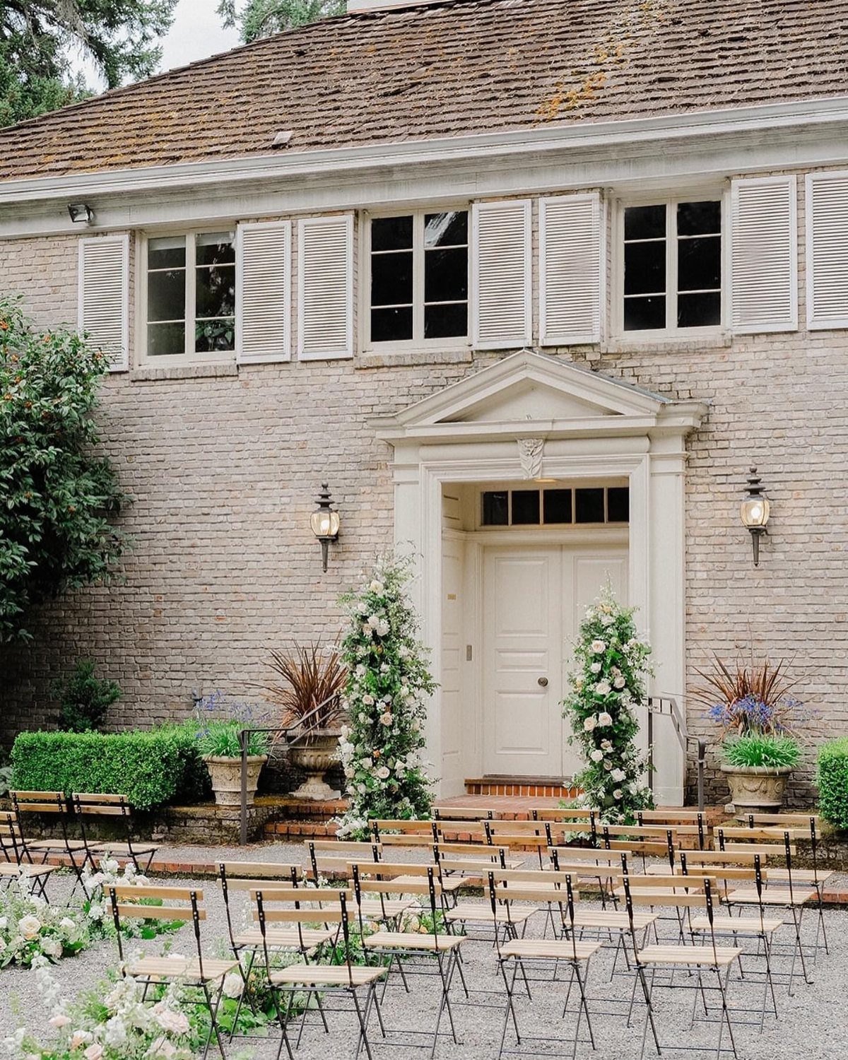 Outdoor wedding ceremony setup with wooden chairs and floral pillars before a brick building.