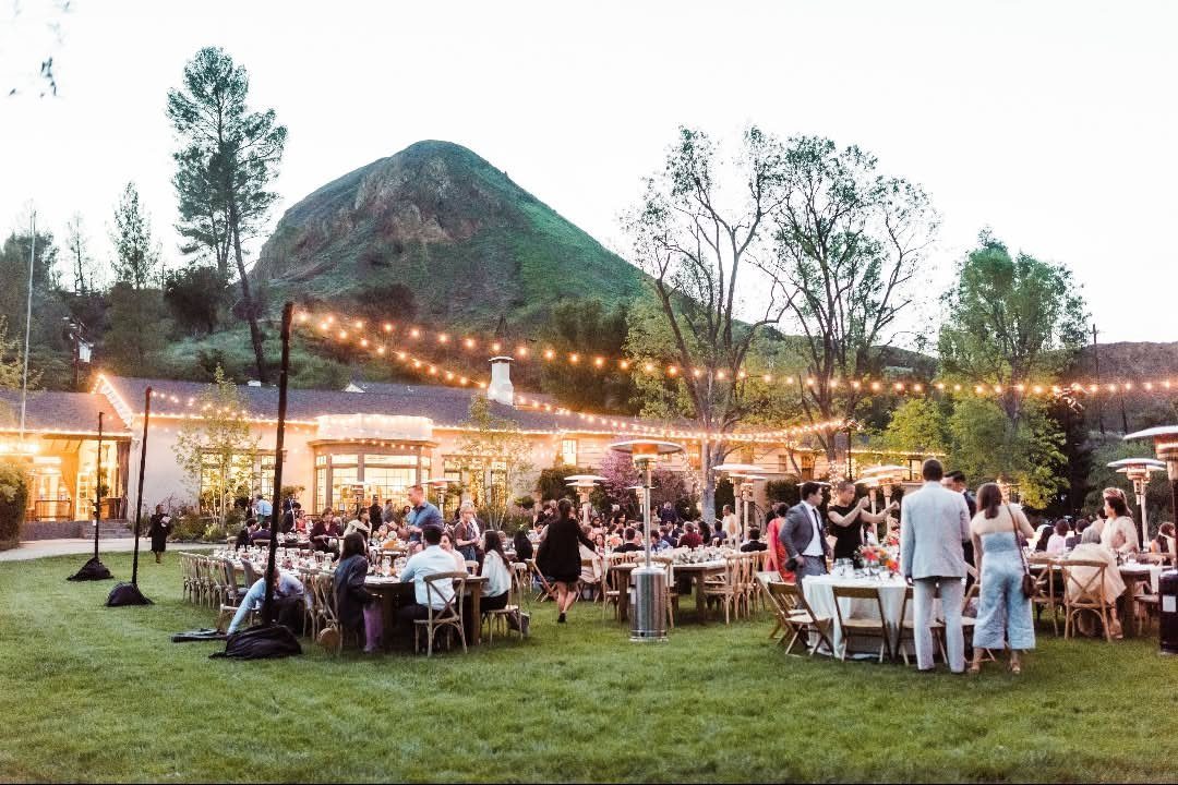 Outdoor wedding reception with guests at tables on a lawn before a large hill.