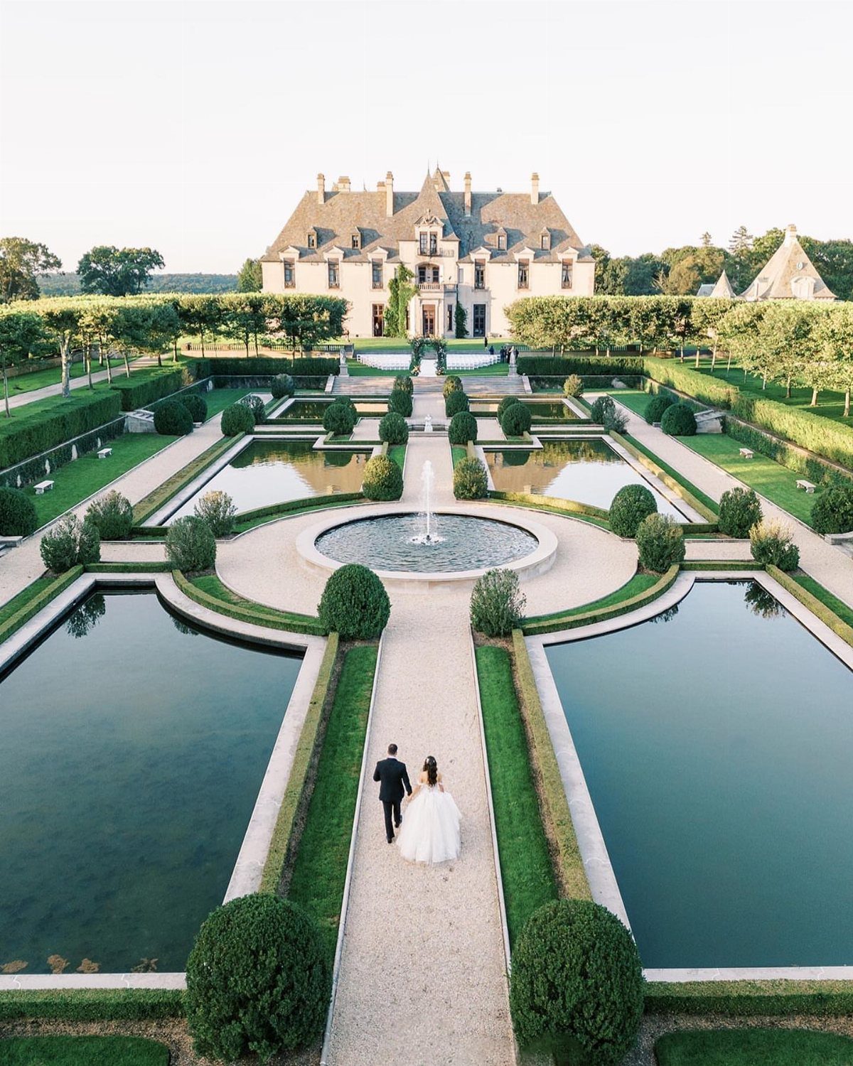 Bride and groom walking through formal gardens toward a grand stone castle estate.