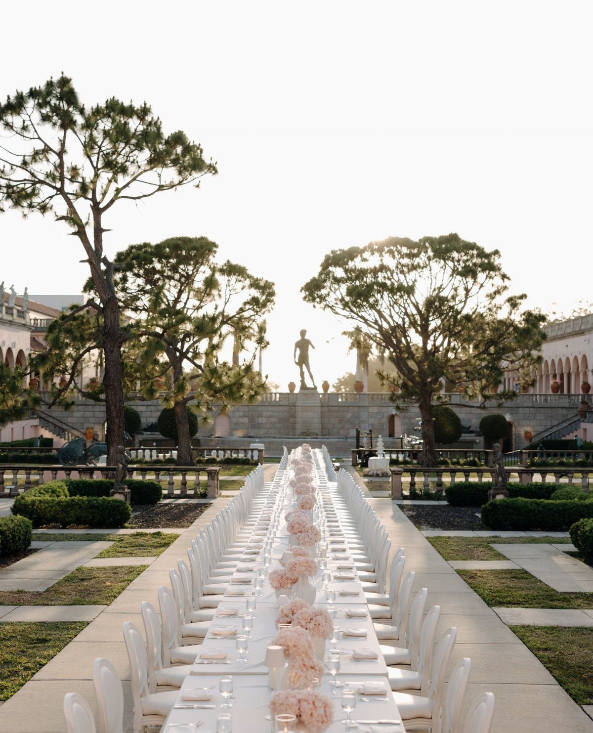 Long white wedding banquet table in a classical courtyard with gardens and statues