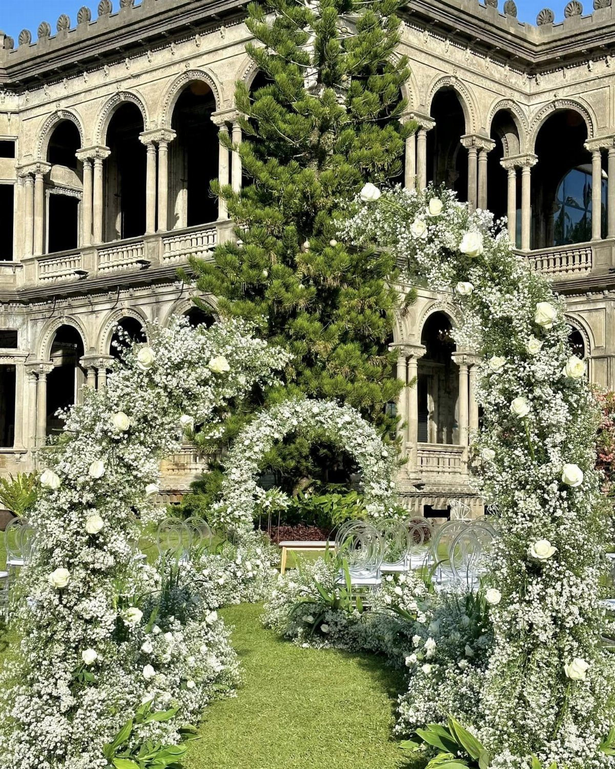 White floral arches leading to a historic stone building with arched colonnades.