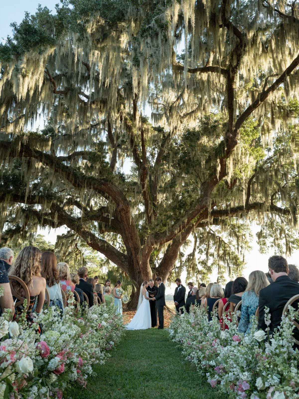 Wedding ceremony under a large oak tree with Spanish moss and floral aisle.