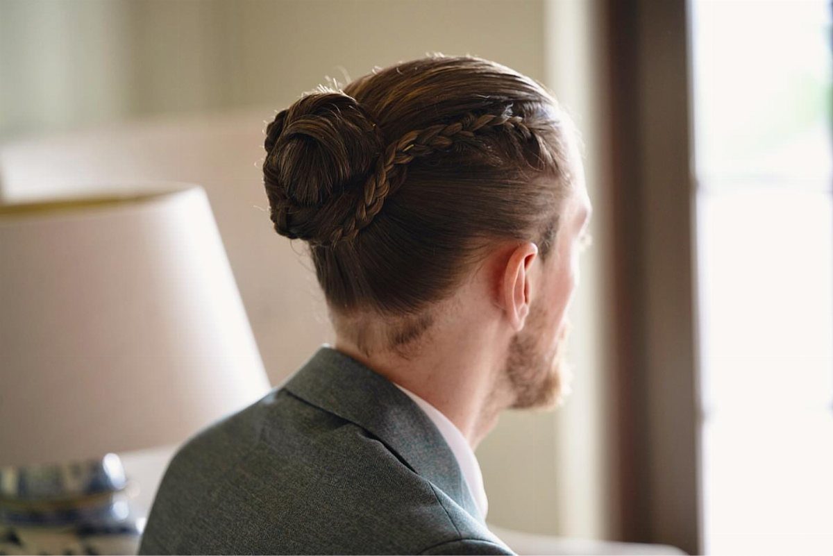 Man with a braided man bun wearing a grey suit jacket seen from behind.
