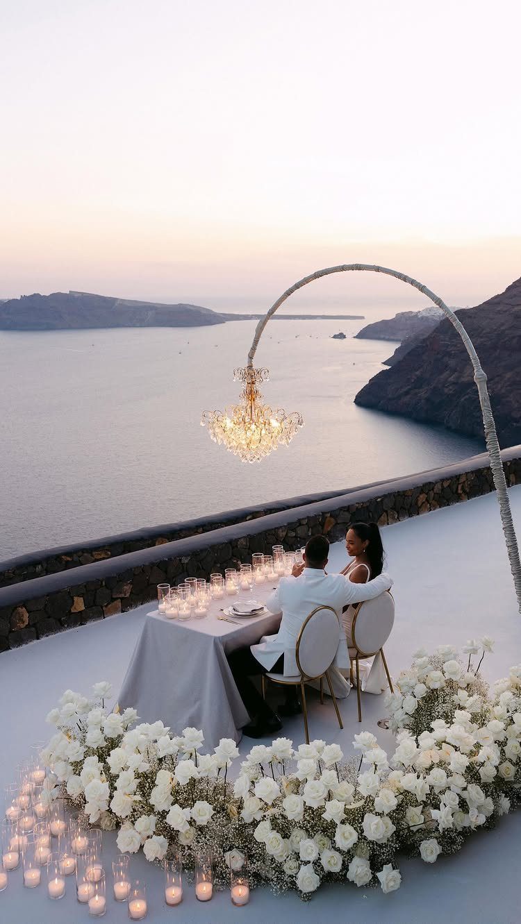Couple in white wedding clothes dining on a terrace with white flowers and chandelier.