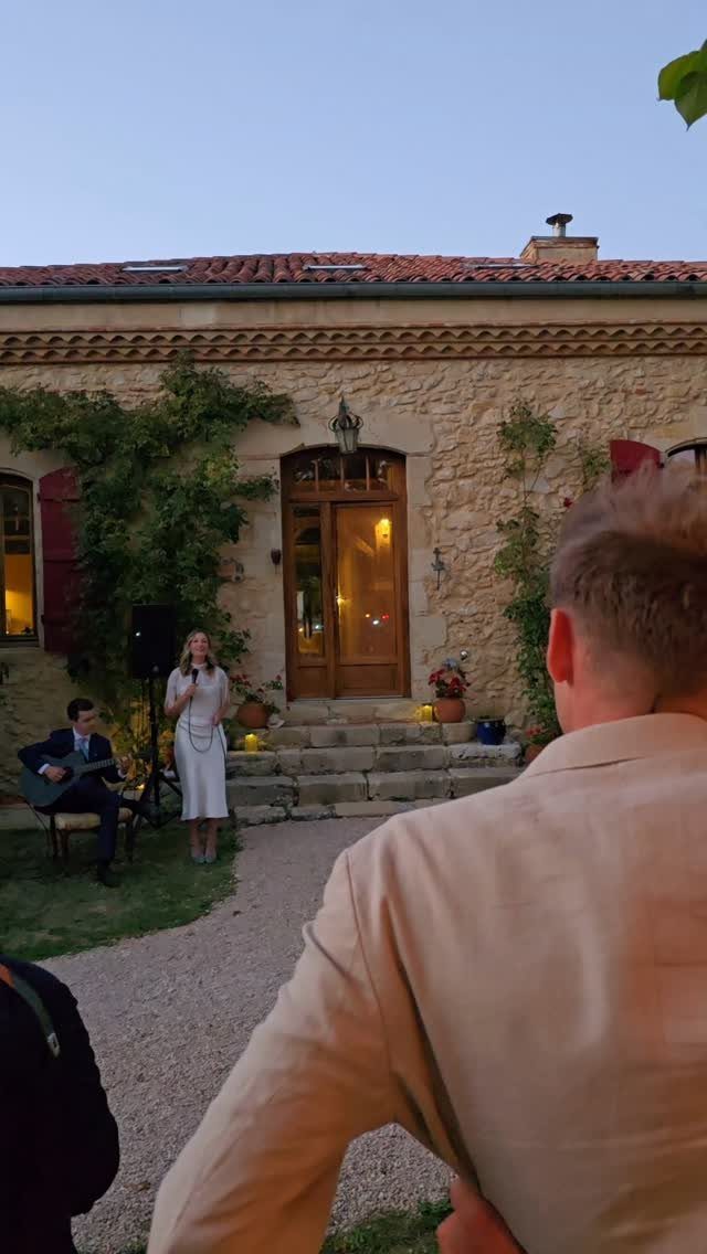 Woman speaking and man playing guitar during a wedding ceremony at a stone house.