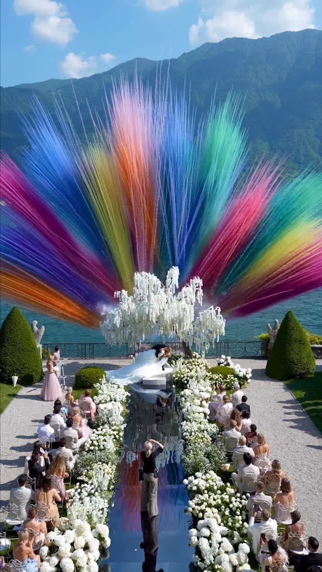Colorful rainbow fabric installation over a white floral wedding ceremony by a lake.