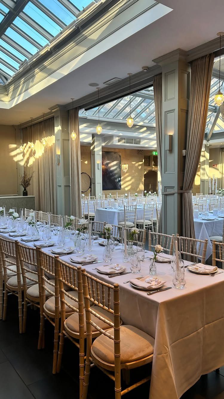 Elegant wedding reception table with gold chairs in a sunlit room with glass ceilings.