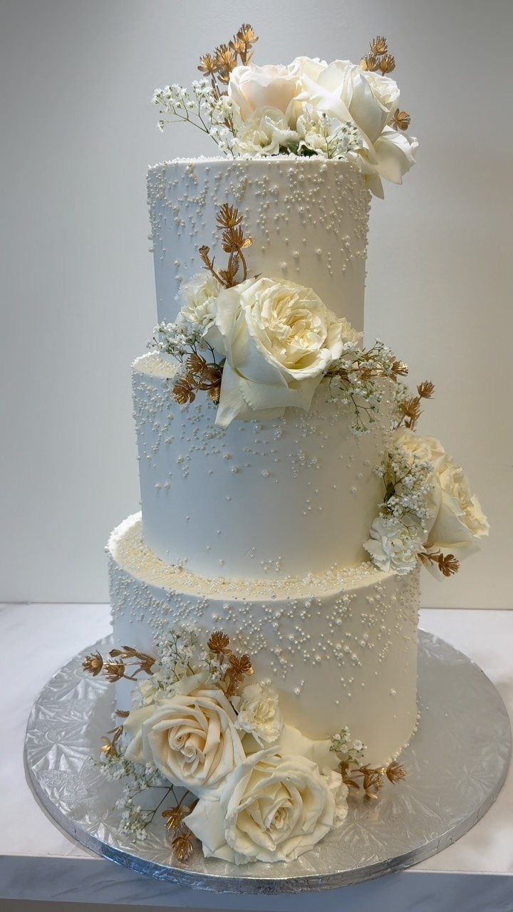 Three-tiered white wedding cake with cream roses and edible pearl decorations on silver platter.