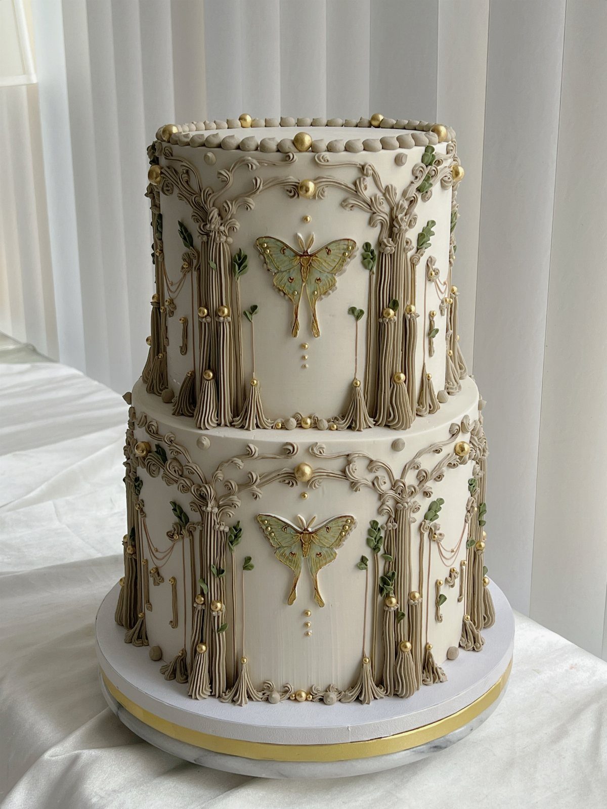 Two-tiered white wedding cake with ornate beige piping, gold pearls, and edible butterflies.