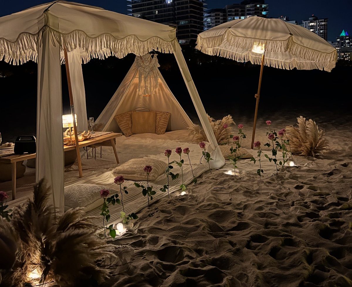 Luxury beach picnic setup with a white canopy, teepee, and candles on the sand.