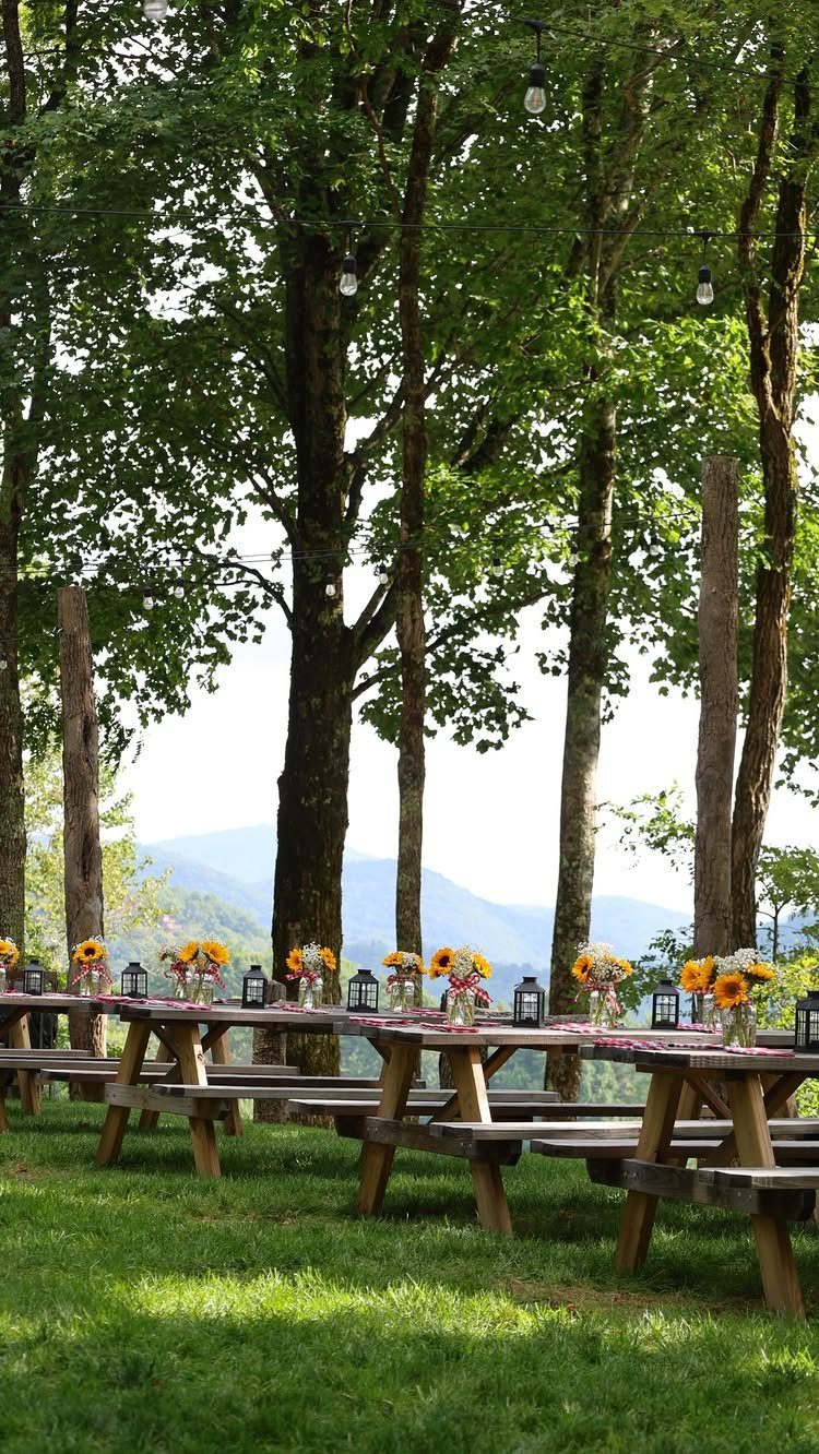 Wooden picnic tables with sunflowers and lanterns set outdoors overlooking blue mountains.