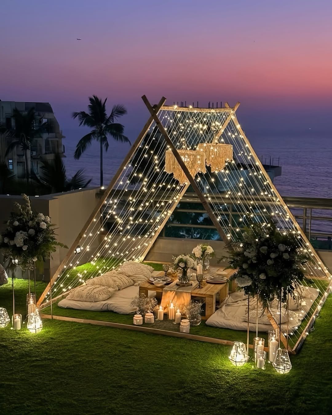 Wooden A-frame structure with string lights and cushions on a lawn at dusk.