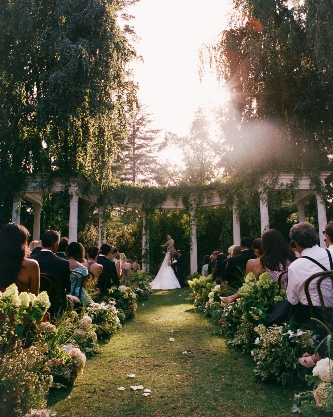 Outdoor wedding ceremony with guests seated along a floral aisle under a white pergola.