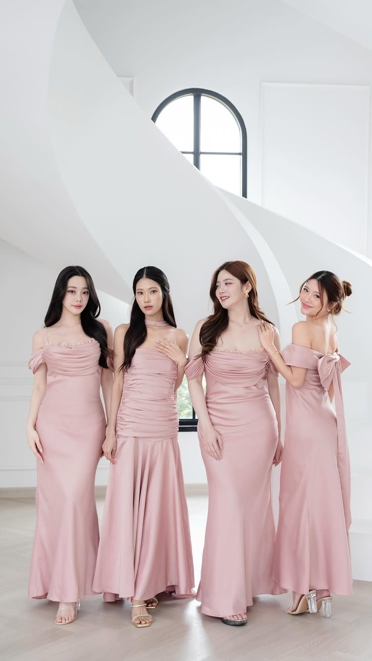 Four women wearing long blush pink satin bridesmaid dresses in a white room.