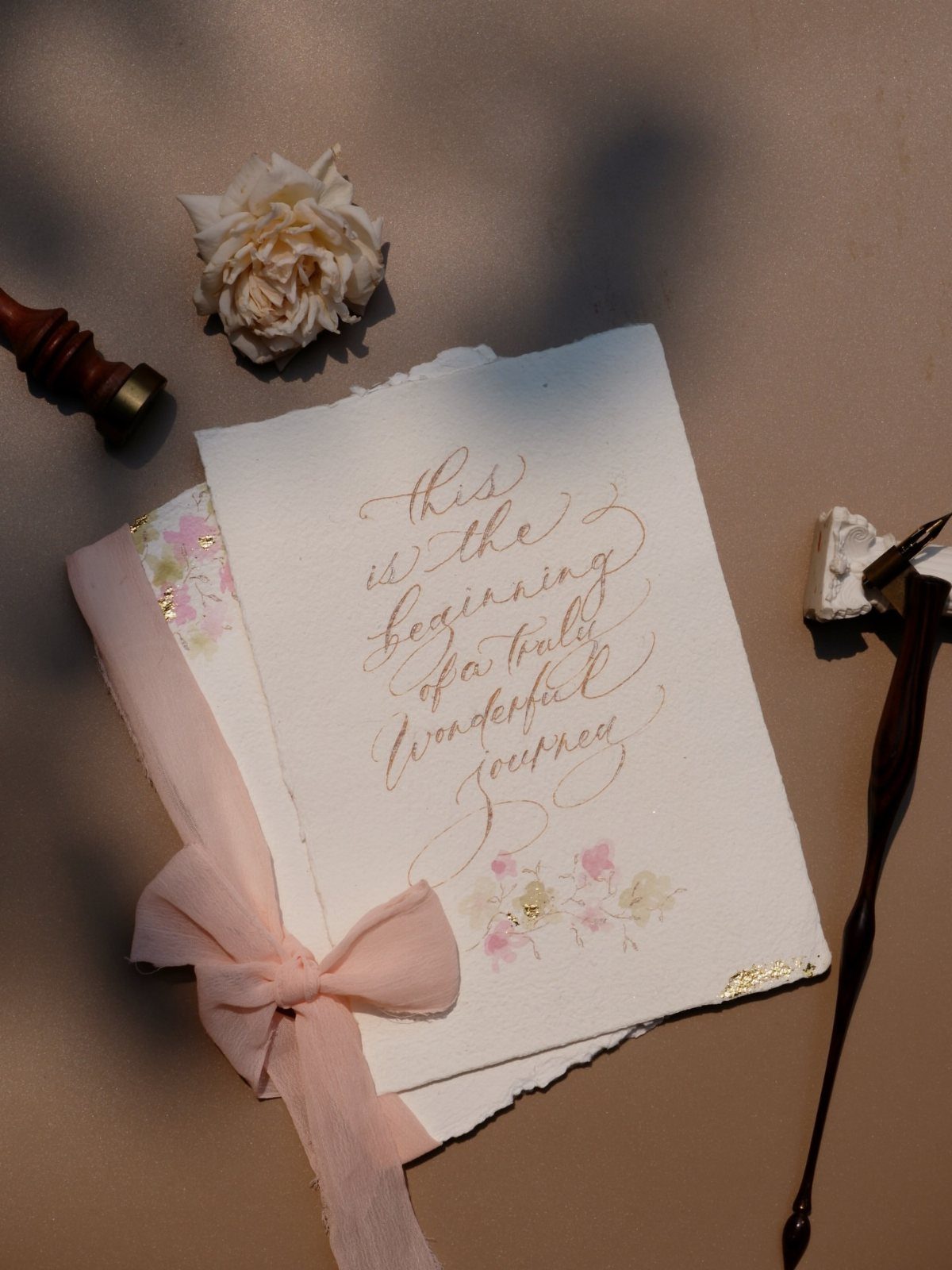 White invitation card with gold calligraphy and pink ribbon next to a white rose.