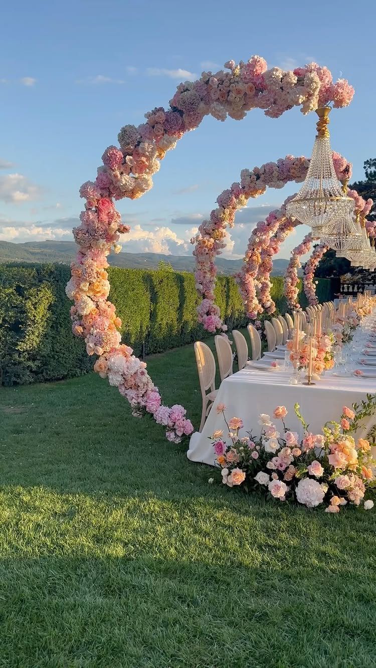 Outdoor dining table with white linens under pink floral arches and a crystal chandelier.