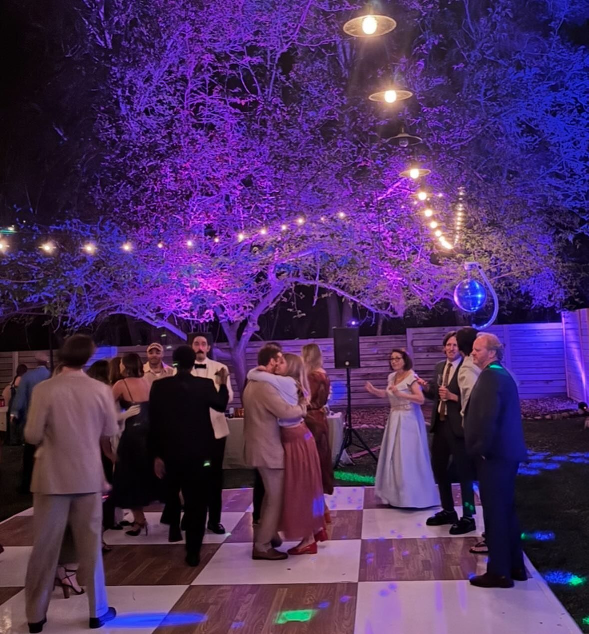 Wedding guests dancing on a checkered floor under a tree with pink ambient lighting.