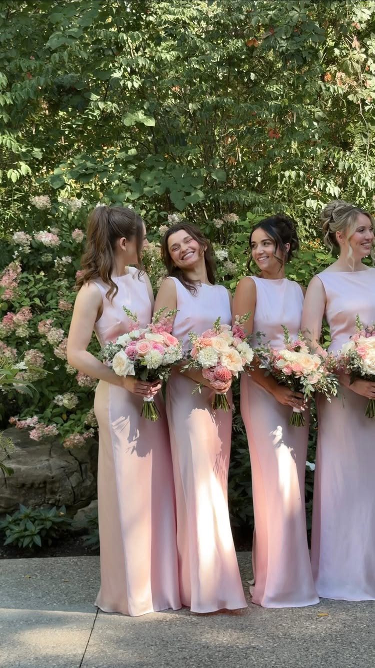 Four bridesmaids in light pink gowns holding floral bouquets against a green garden backdrop.