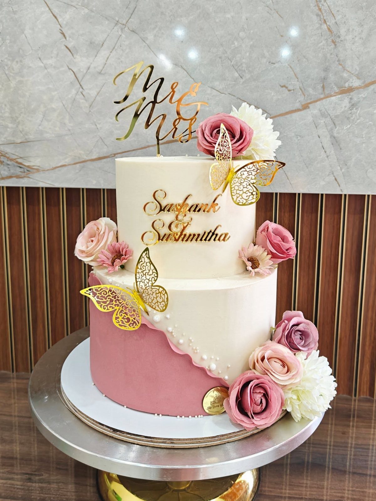 Two-tier white and pink wedding cake decorated with roses and gold butterflies.