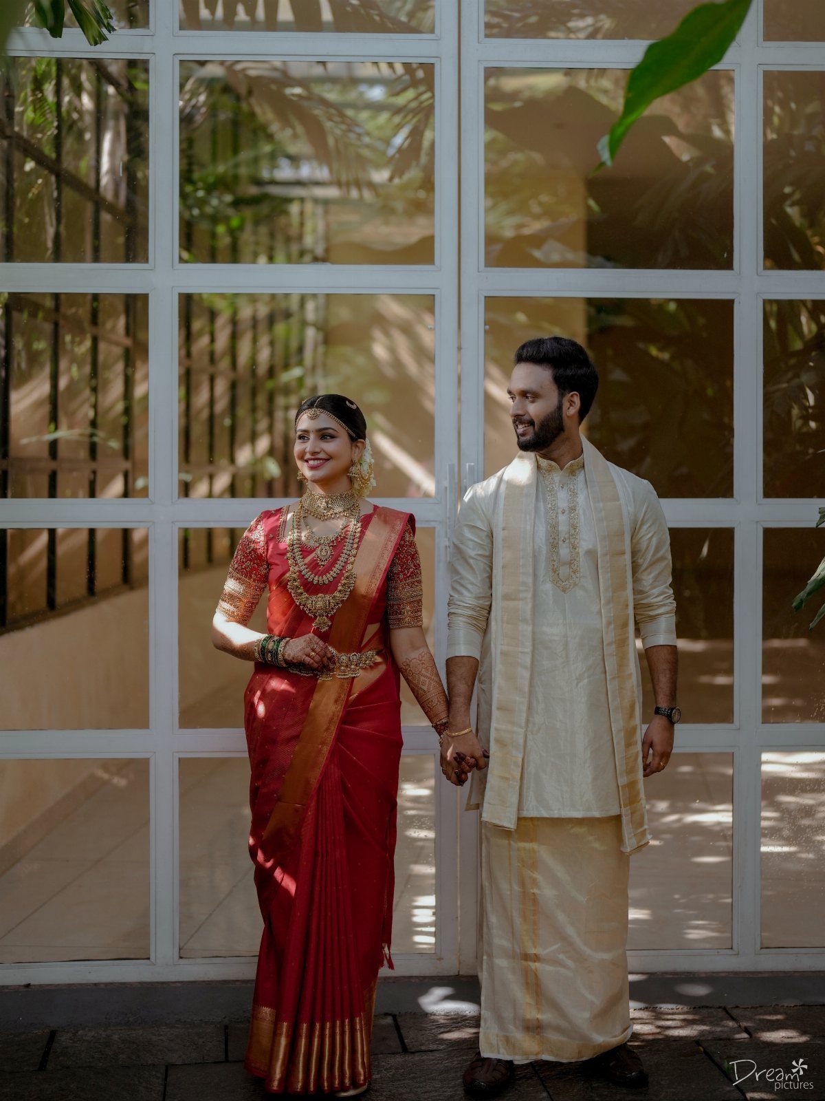 12 Pre Wedding Photoshoot Outfit Ideas for Couples 3 Couple in traditional Indian wedding attire posing before a white grid window.