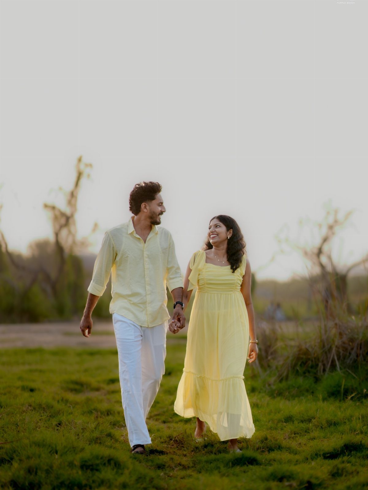 12 Pre Wedding Photoshoot Outfit Ideas for Couples 5 Couple in matching pale yellow outfits walking through a grassy field at sunset.