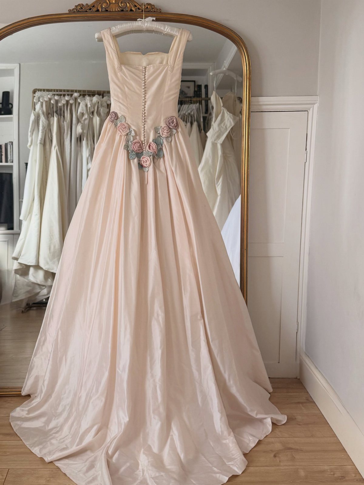 Blush satin wedding dress with floral waist appliques hanging before a gold mirror.