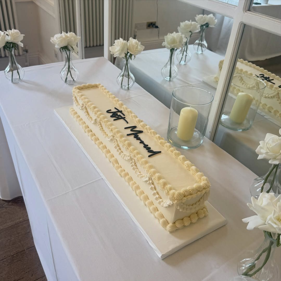 Rectangular white wedding cake with cursive writing and white roses on a table.