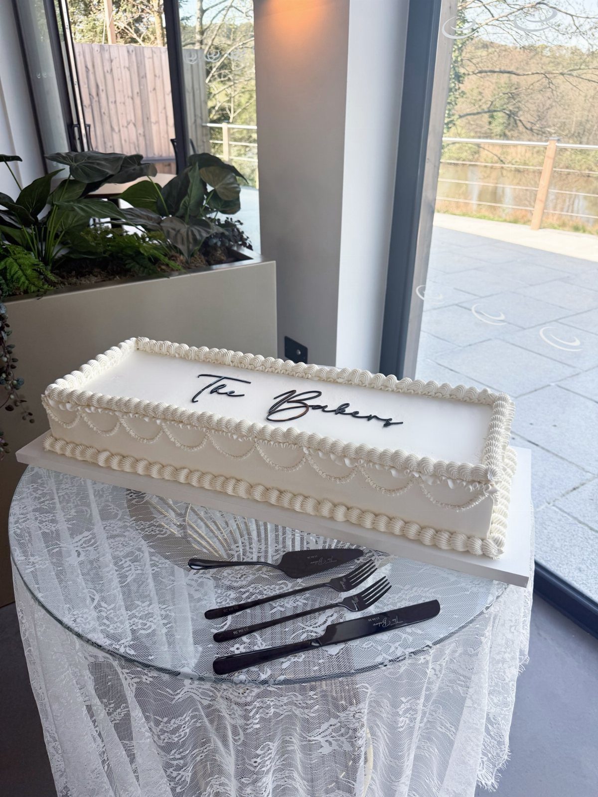 Rectangular white buttercream cake with piped borders and black script on a glass table.