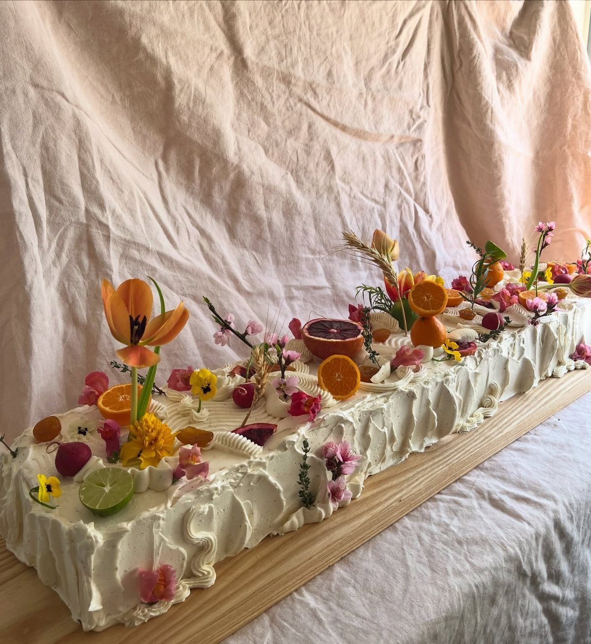 Rectangular white frosted cake decorated with fresh citrus fruits and colorful spring flowers.