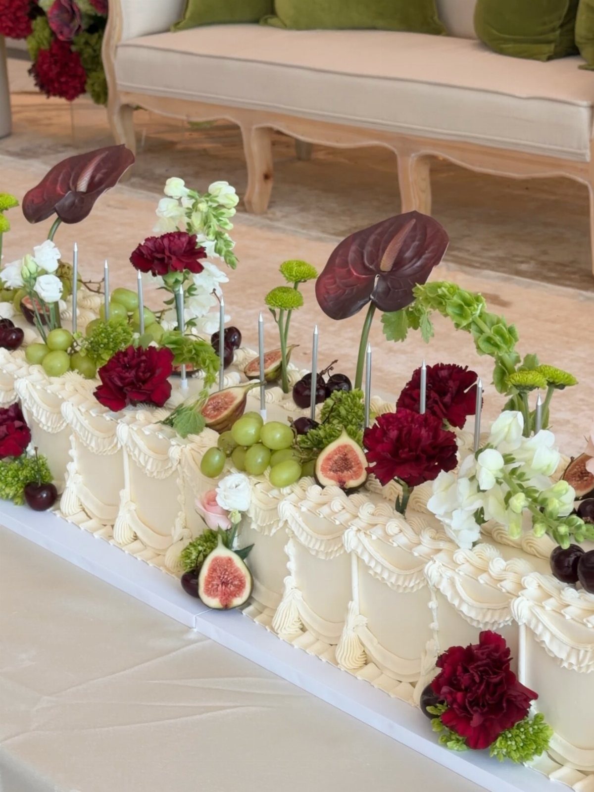 Long rectangular white cake decorated with red flowers, green grapes, figs, and greenery.