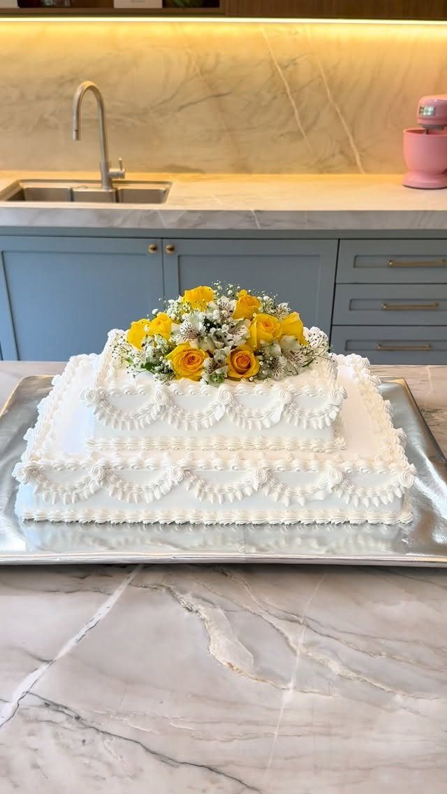 Two-tier white rectangular cake with scalloped piping and yellow roses on marble counter.