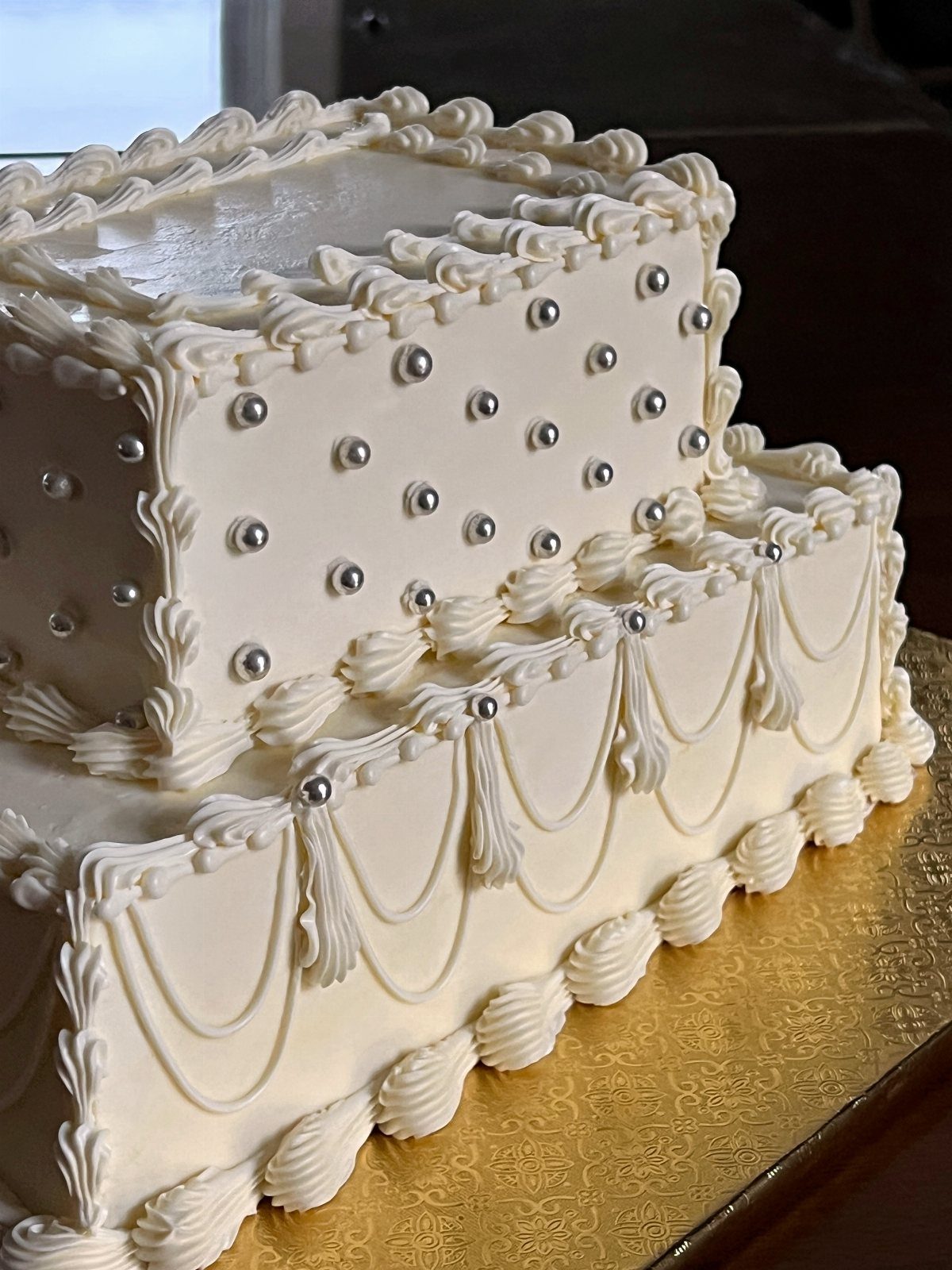Two-tier rectangular white wedding cake with silver pearls and ornate frosting on gold base